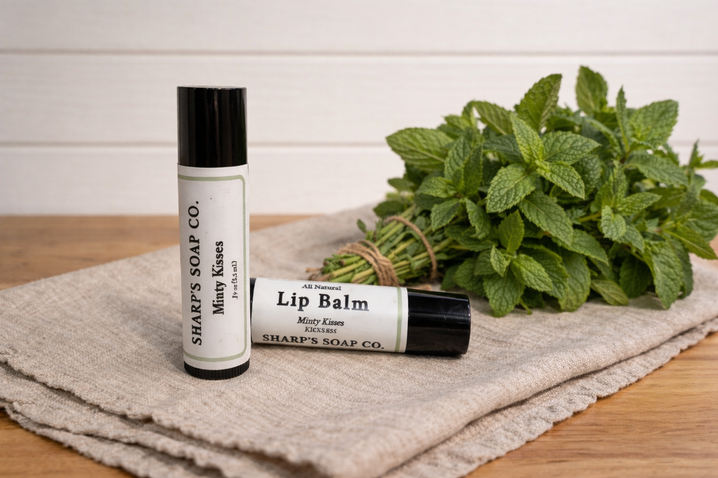Two lip balm tubes and a bundle of mint leaves on a textured cloth with a white background.