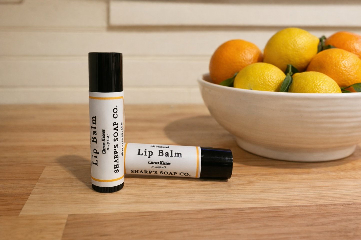 Two lip balm tubes from Sharp's Soap Co. on a wooden surface with a bowl of lemons and oranges in the background.