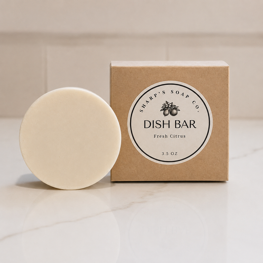 A round off-white dish soap bar standing in front of its kraft paper box, which features a white circular label reading 'SHARP'S SOAP CO. DISH BAR Fresh Citrus 3.5 oz'
