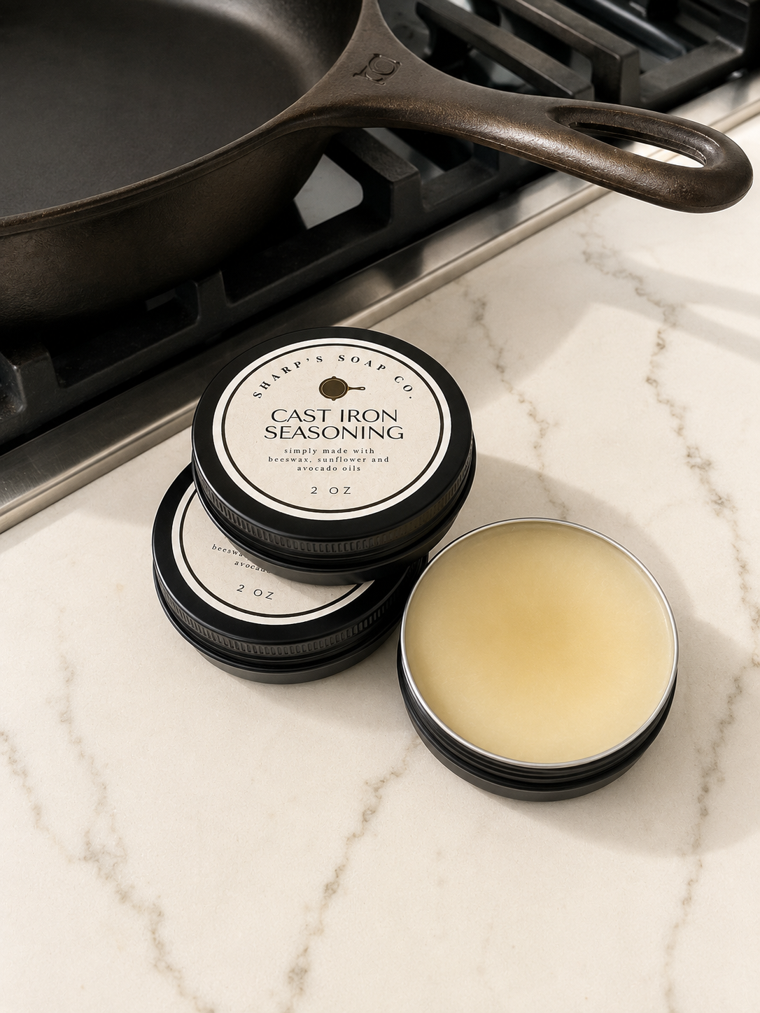 Three small round black tins of Sharp's Soap Co. Cast Iron Seasoning are arranged on a white marble countertop with a cast iron skillet in the background.